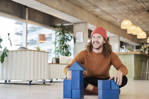 Cheerful man playing with toy block while sitting on floor in living room at home - FMKF06721