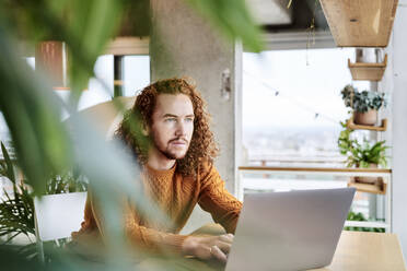 Hipster man using laptop looking away while sitting at home - FMKF06728