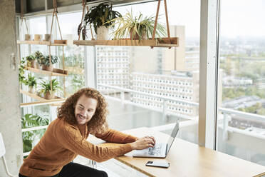 Redhead man looking over shoulder while using laptop sitting at home - FMKF06732