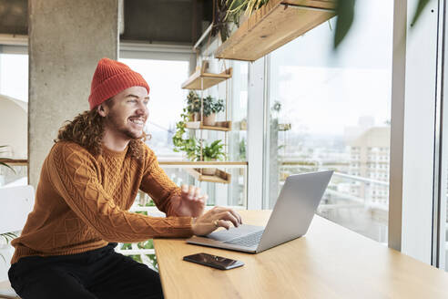 Smiling man wearing knit hat using laptop sitting at home - FMKF06733