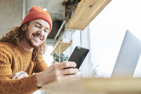 Smiling hipster man using smart phone with laptop sitting at home - FMKF06735