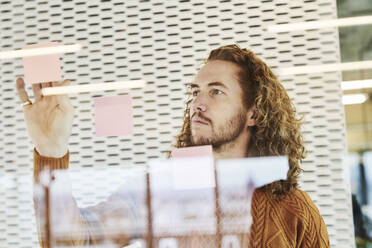 Hipster man sticking sticky notes on glass material at home - FMKF06737