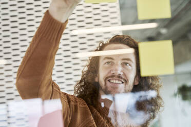 Smiling man sticking adhesive notes on glass material at home - FMKF06739