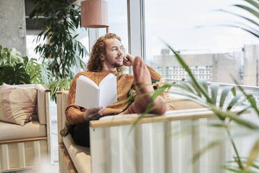 Nachdenklicher Mann, der durch das Fenster schaut und ein Buch hält, während er zu Hause auf dem Sofa sitzt - FMKF06745