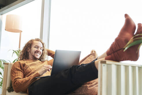 Smiling man using laptop while lying on sofa at home - FMKF06746