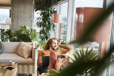 Man with laptop lying down on sofa looking through window at home - FMKF06748