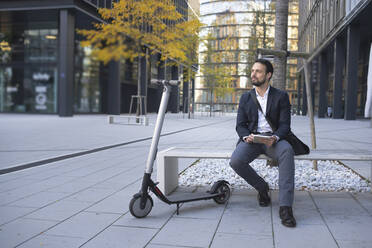 Businessman with digital tablet sitting by e-scooter on bench in city - HMEF01137