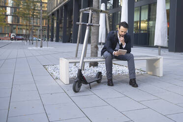 Businessman using digital tablet while sitting by e-scooter on bench in city - HMEF01138