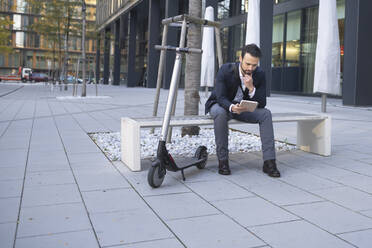 Geschäftsmann, der ein digitales Tablet benutzt, während er mit einem E-Scooter auf einer Bank in der Stadt sitzt - HMEF01138