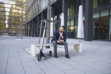 Businessman with digital tablet looking away while sitting by e-scooter on bench in city - HMEF01139