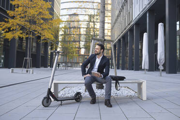 Unternehmer mit digitalem Tablet auf einer Bank in der Stadt neben einem E-Scooter sitzend - HMEF01140