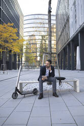 Entrepreneur with digital tablet sitting by electric scooter on bench in city - HMEF01141