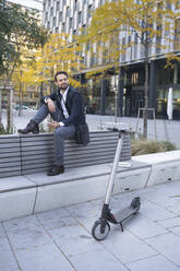 Smiling entrepreneur with in-ear headphones sitting on retaining wall in city - HMEF01144