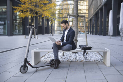 Smiling businessman using laptop while sitting by push scooter on bench in city - HMEF01145