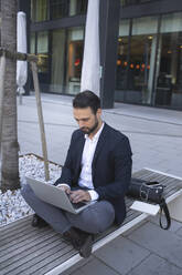 Confident businessman using laptop while sitting on bench in city - HMEF01146