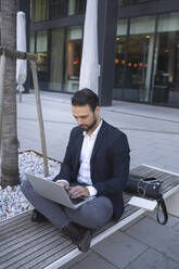 Selbstbewusster Geschäftsmann, der einen Laptop benutzt und auf einer Bank in der Stadt sitzt - HMEF01146