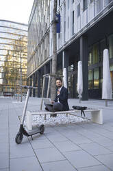 Smiling businessman using laptop by electric push scooter on bench in city - HMEF01147