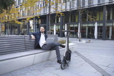 Businessman looking away while using laptop by push scooter in city - HMEF01149