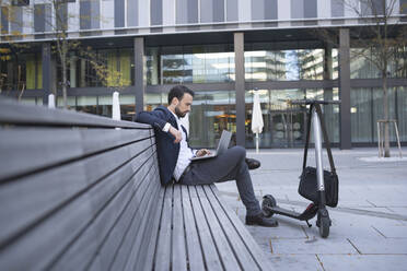 Businessman looking at laptop while sitting on retaining wall by push scooter in city - HMEF01151