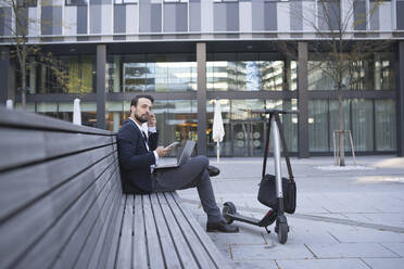 Businessman using in-ear headphones while sitting on retaining wall in city - HMEF01152