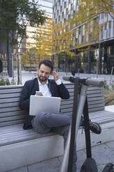 Businessman using in-ear headphones on retaining wall in city - HMEF01153