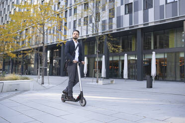 Businessman riding on electric scooter against building in city - HMEF01154