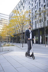 Male business person riding on electric scooter against building in city - HMEF01155
