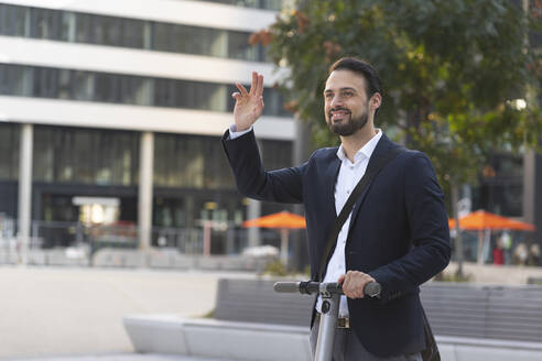 Lächelnder Geschäftsmann winkt auf einem Elektroroller in der Stadt - HMEF01159