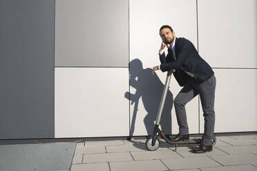 Confident entrepreneur looking away while leaning on electric push scooter in city - HMEF01163