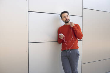 Contemplating entrepreneur with in-ear headphones standing against wall - HMEF01169