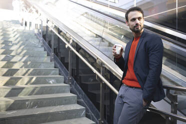 Selbstbewusster Unternehmer trinkt Kaffee, während er auf einer Treppe in der Stadt steht - HMEF01173