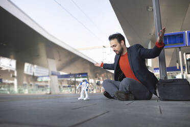 Confident entrepreneur with arms outstretched looking at robot while sitting on railroad platform - HMEF01178
