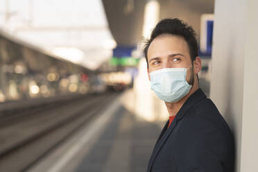 Male entrepreneur looking away while wearing protective mask on railroad platform - HMEF01180