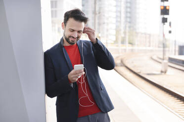 Smiling entrepreneur with in-ear headphones standing on railroad platform - HMEF01187