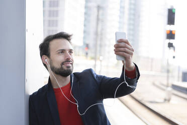 Smiling entrepreneur with in-ear headphones standing by wall on railroad platform - HMEF01188