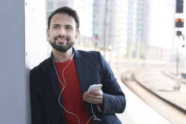Smiling entrepreneur with in-ear headphones waiting on railroad platform - HMEF01189