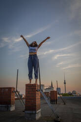 Happy woman with arms raised standing on rooftop against sky during sunset - NGF00716