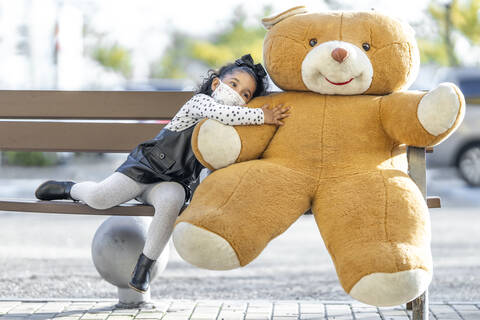 Girl wearing face mask embracing teddy bear while sitting on bench stock photo