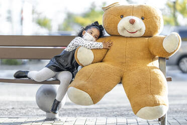 Mädchen mit Gesichtsmaske umarmt Teddybär, während sie auf einer Bank sitzt - GGGF00183