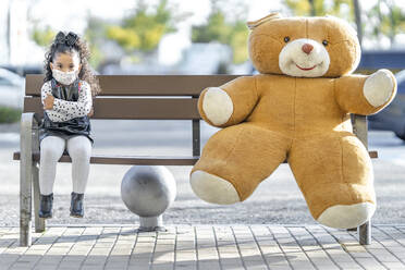 Mädchen mit Gesichtsmaske sitzt mit Teddybär in sozialer Distanz auf einer Bank - GGGF00185