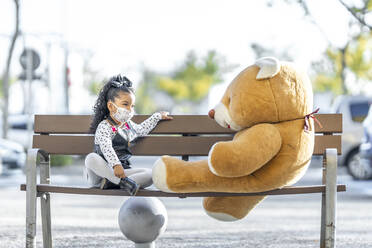 Mädchen mit Gesichtsmaske, das mit einem Teddybären spielt, während es in sozialer Distanz auf einer Bank sitzt - GGGF00188