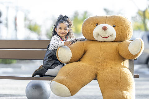 Smiling girl sitting with teddy bear on bench stock photo