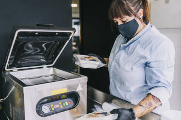 Female chef using airtight packing machinery while holding take out food at restaurant during pandemic - DGOF01685