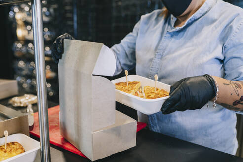 Midsection of female chef putting fresh food in cardboard box for take out at kitchen counter - DGOF01701