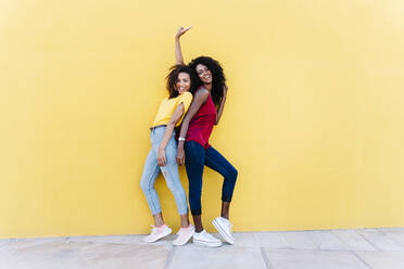 Cheerful girlfriends enjoying while standing back to back by yellow wall - RDGF00212