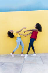 Cheerful women holding hands while pointing at each other by yellow wall - RDGF00213