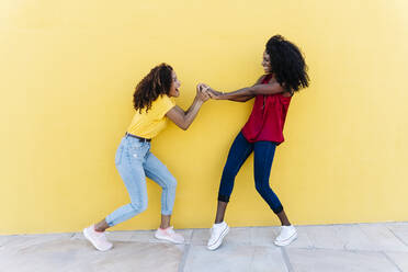 Lesbisches Paar hält sich lachend an der gelben Wand die Hände - RDGF00214