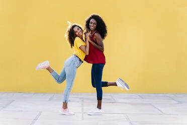 Happy lesbian couple holding hands while standing on one leg against yellow wall  - RDGF00219