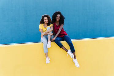Smiling young female homosexual couple sitting on yellow retaining wall - RDGF00225