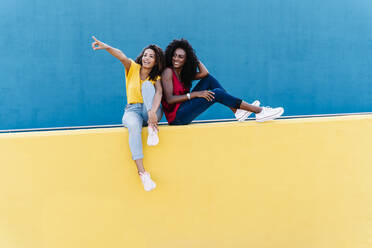 Young woman pointing to smiling friend while sitting on yellow retaining wall - RDGF00226